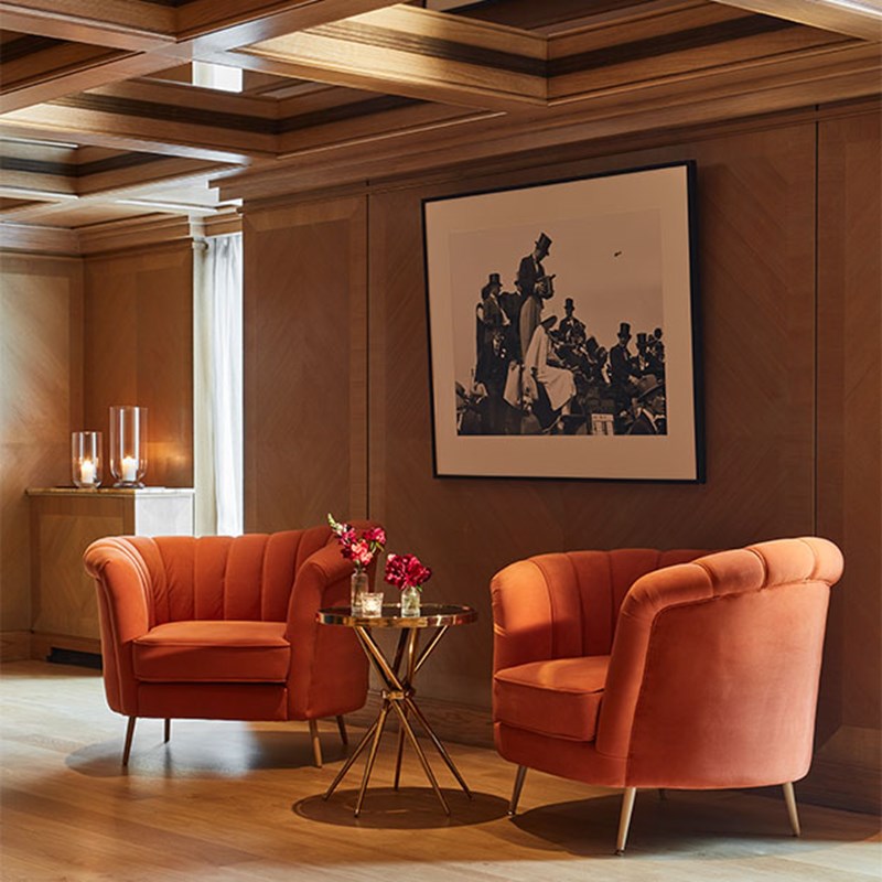 Two plush orange chairs create a cozy seating area. A round gold glass-topped table with red flowers and a lit candle sits between the chairs. A framed black and white photograph hands on the wood-paneled walls.