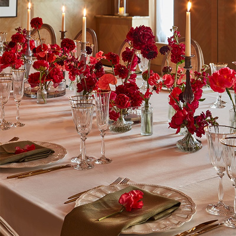 An elegantly set dinner table features white tablecloths, crystal glassware, lit candles, red floral arrangements and gold flatware. Each place setting includes a green napkin with a single red flower.