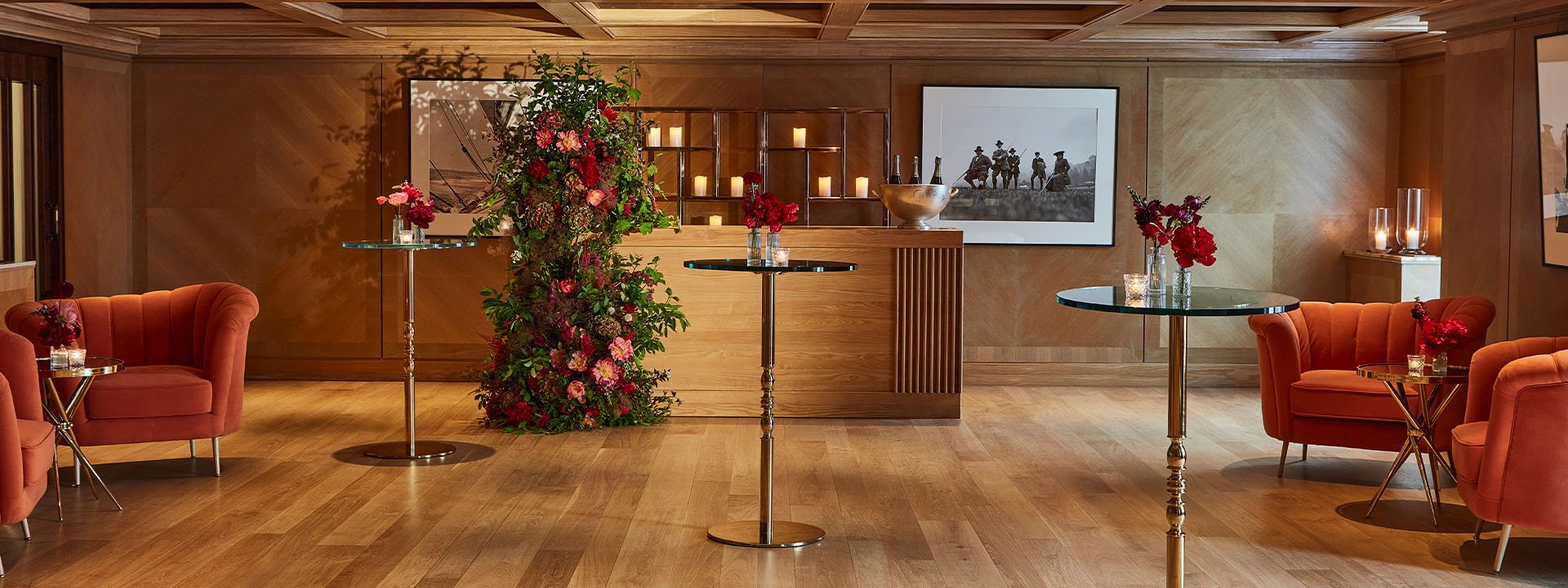 A warmly lit room features a wooden bar with candles and floral arrangements. Tall gold glass-topped tables are positioned throughout, and plush orange chairs create a cozy seating area. Framed black and white photographs hang on the wood-paneled walls.