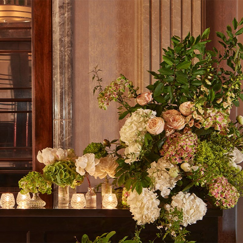 A big bouquet of white and pink flowers with greenery is sitting atop of a wooden bar with vases of flowers and candles lit on its left.