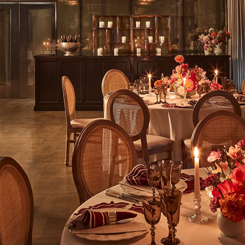 A luxurious dining room is set for an event featuring two round tables with tablecloths, candles, a vase filled with pink flowers and stylish chairs with wicker backs. To the back is a wooden bar with candles and bottles of champagne.