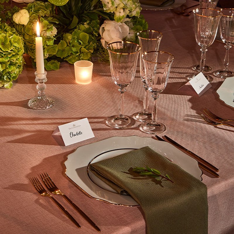 An elegantly set dinner table features a white plate with a green napkin, gold flatware, crystal glasses, and a place card labeled