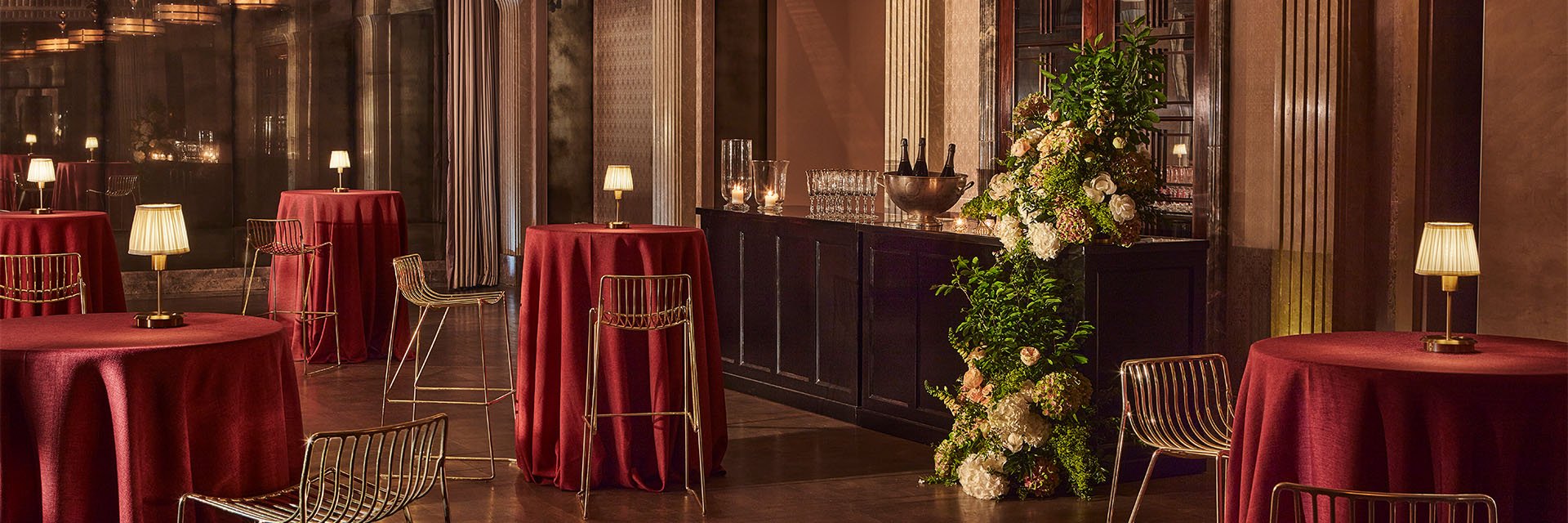 High tables draped in rich red tablecloths and topped with elegant lamps create a sophisticated setting. The dark wood bar area is adorned with floral arrangements, adding a touch of freshness and charm to the luxurious ambiance.