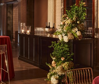 A stylish bar area with floral decorations and glassware is set up beside a small round table with a red tablecloth and a gold chair. The room features warm lighting and elegant décor.