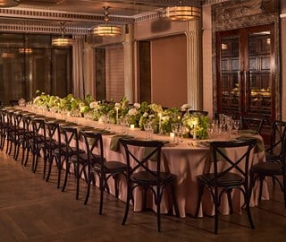 A luxurious dining room is set for an events, featuring a long oval table with a tablecloth, floral centrepieces and candlelight, surrounded by wooden chairs.
