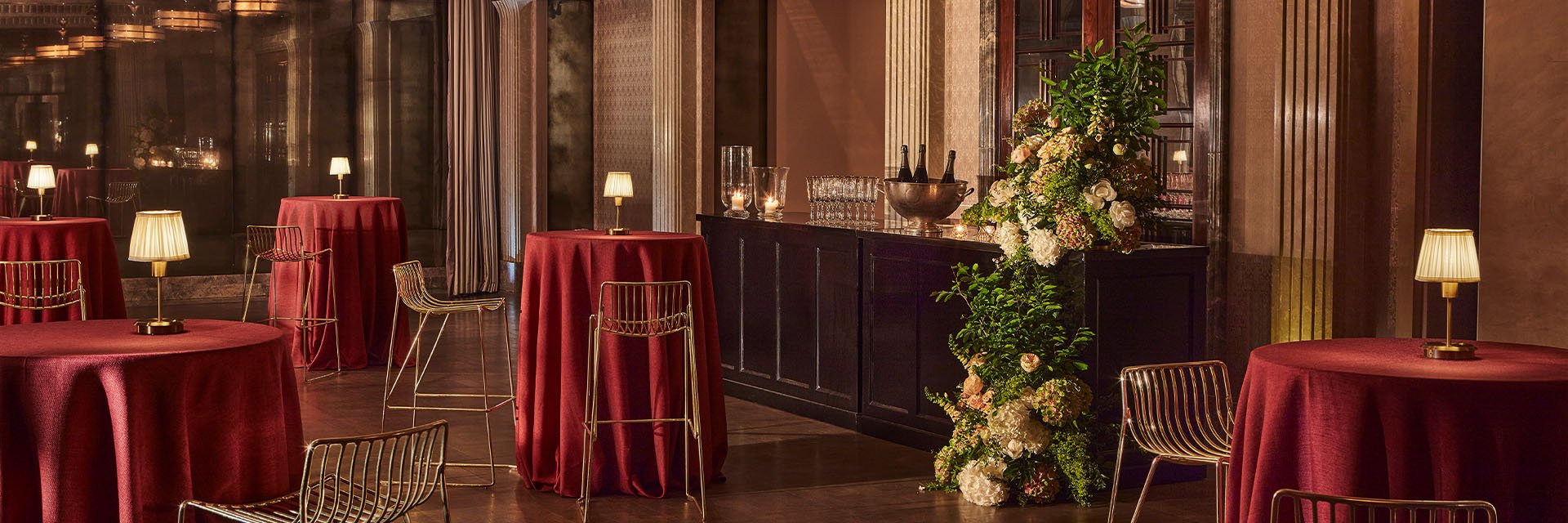 High tables draped in rich red tablecloths and topped with elegant lamps create a sophisticated setting. The dark wood bar area is adorned with floral arrangements, adding a touch of freshness and charm to the luxurious ambiance.