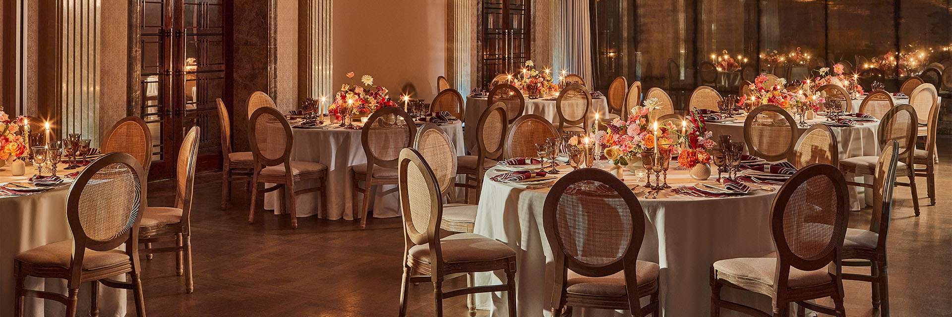 Luxurious room elegantly laid up with round tables, with white table clothes, centre piece flowers and wicker backed chairs.