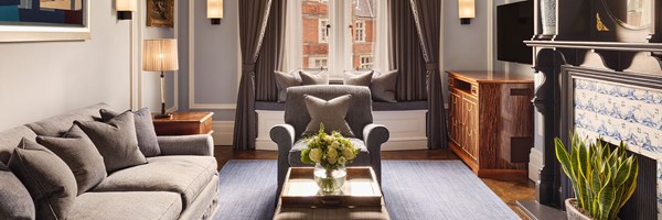 Elegant living room featuring a grey sofa and armchair with scatter cushions, a window seat with plush pillows, and a central coffee table topped with fresh flowers, framed by tall windows with dark drapes and views of redbrick buildings.