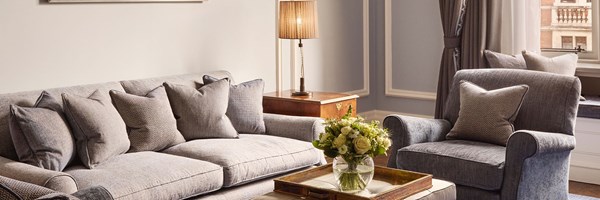 Detail view of the Carlos Suite living area showing a textured grey sofa and matching armchair, both with coordinated cushions, a side table with a lamp, and a flower arrangement on a wooden tray.