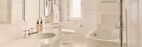 Light-filled bathroom with double vanity sinks, a freestanding bathtub, and polished chrome fixtures, featuring mirrored cabinets, a window with frosted glass, and white walls throughout.