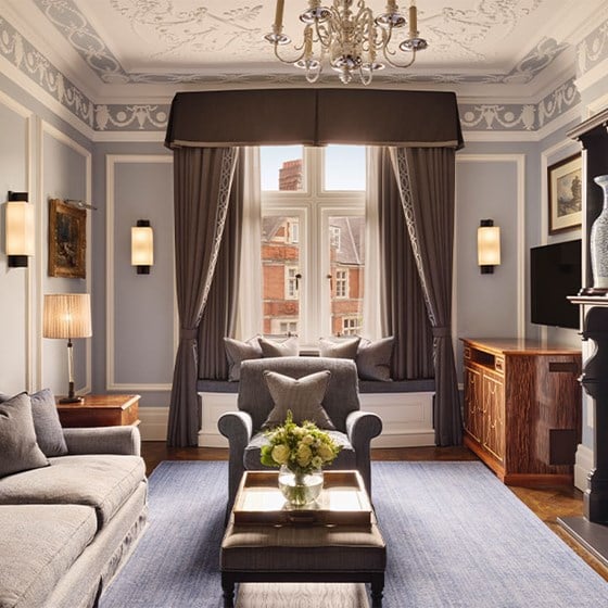 Wide shot of the Carlos Suite living room with detailed ceiling mouldings, chandelier, and elegant decor including a grey sofa, armchair, dark fireplace, window seat, and classic wood furnishings.