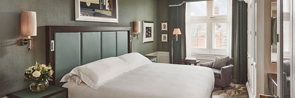 Double bed with white bedding, green eadboard, green bed side tables with a vase of flowers. Beside the bed there is an arm chair and coffee table with books.