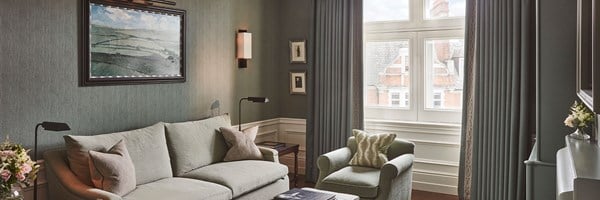 Coburg Suite: Living area of suite with sofa and cushions on the left, and a painting on the wall. A coffee table with books in front of the sofa, and a chair with a green cushion. The colours are shades of green.