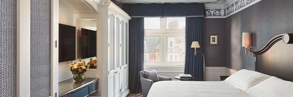 On the left: cupboards and a television, and on the right a double bed with a wooden headboard. Window at the back framed with blue curtains.