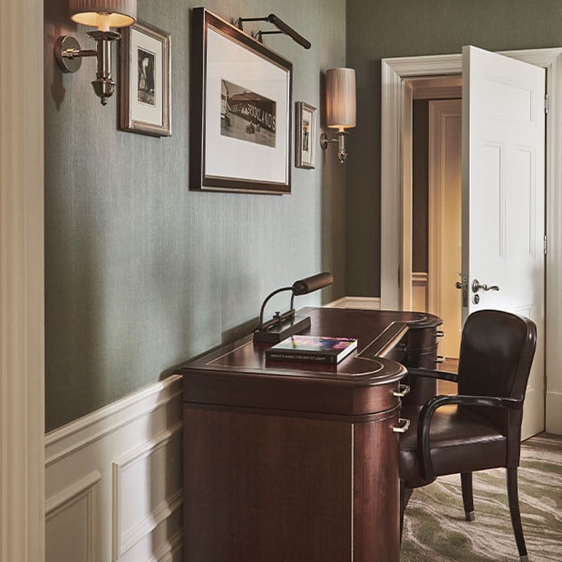 Side view of wooden desk with brown leather chair tucked behind it. The desk faces the wall, and there are three pictures hung up, the middle one is large and rectangle, and the pictures either side are smaller and square.