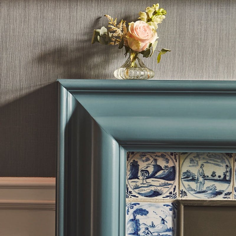 View of corner of fire place, with blue and white tiles depicting stories. On the mantle piece is a small vase of flowers.