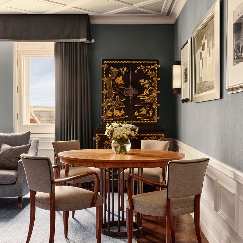 Dining area in Suite 205, featuring mid-century style armchairs, a round wooden table, and a lacquered Chinoiserie cabinet set against soft blue walls.