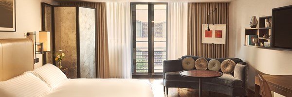 Bright studio bedroom with tall windows, a blue curved sofa and round table beside neutral drapes.