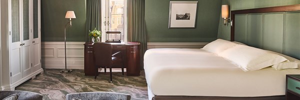 A spacious bedroom with a large white bed, green walls and a desk by the window overlooking townhouse rooftops.