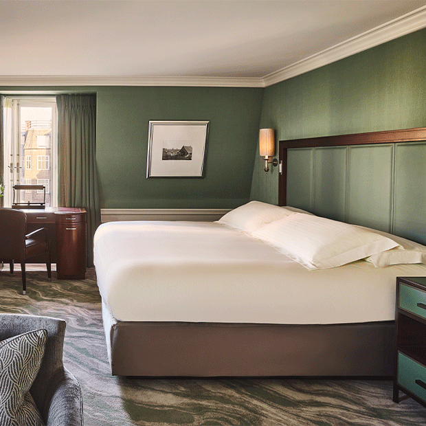 A green-toned bedroom with a large white bed, desk by the window and soft patterned carpet.