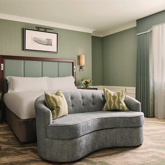 A serene hotel room featuring a plush king-sized bed with a soft blue tufted sofa at its foot, adorned with elegant green cushions and framed by soft, muted green walls.