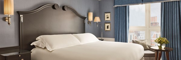Elegant bedroom with a dark wood headboard, king bed dressed in white linens, blue walls and curtains, and a seating area by the window with a round side table and flowers.