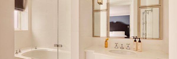 Bright bathroom with a white vanity and mirror, large bathtub with glass shower screen, and chrome fixtures, with the bedroom visible in the reflection.