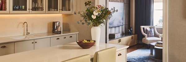 A bright kitchen with pale cabinetry, marble countertops, and glass-fronted cupboards, featuring a vase of flowers and fruit bowl on the island, leading into a sitting area with an armchair and blue curtains.