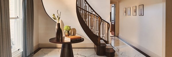 A marble-floored hallway with a curved wooden staircase and geometric balustrade, next to a round table decorated with books and sculptural vases beneath tall windows with patterned curtains.