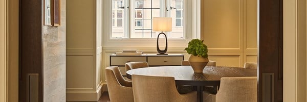 A softly lit dining room with a round wooden table surrounded by upholstered chairs, a large window with a table lamp, and a simple arrangement of green hydrangeas in a ceramic vase.