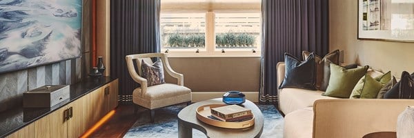 A modern sitting room with a cream sofa and green and navy cushions, a beige armchair by a window with dark blue curtains, and an abstract artwork above a low black sideboard.