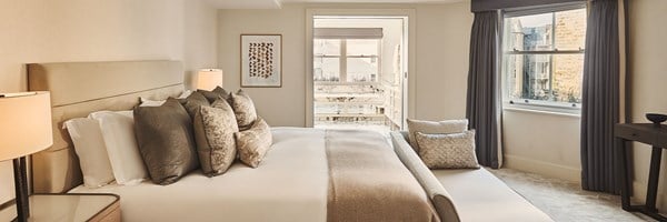 Spacious bedroom with a king-size bed dressed in soft neutral tones, large windows with grey curtains, and an adjoining marble bathroom visible through open doors.
