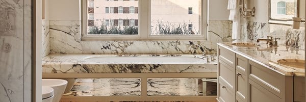 Elegant marble bathroom with a built-in bathtub beneath a large window, double sinks with chrome fixtures, and light cabinetry for a polished, luxurious look.