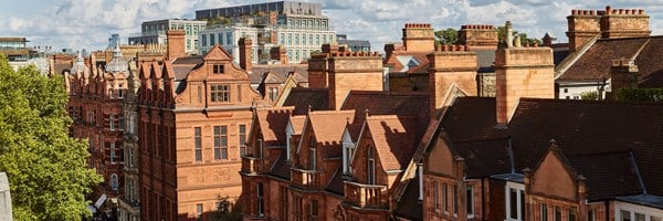 A rooftop view over red-brick Edwardian buildings in Mayfair, London, under a bright blue sky with scattered clouds.