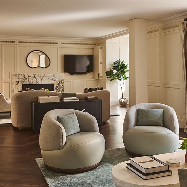 A spacious living room with curved armchairs, a sofa facing a marble fireplace and wall-mounted television, decorated in beige and soft green tones with wooden floors and panelled walls.