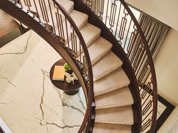 An elegant curved staircase with a dark wooden handrail and geometric balustrade descending to a marble-floored hallway with a round table and sculptural decor beneath.