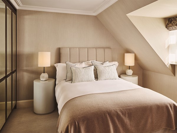 A softly lit bedroom with a double bed dressed in white linens and beige throw, flanked by two round bedside tables with matching lamps, under a sloped ceiling and neutral-toned walls.