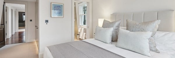 Softly lit bedroom with king bed, taupe and silver bedding, fitted wardrobes, and open door to hallway and en suite.