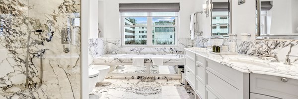 Luxurious bathroom in white marble with double sinks, walk-in shower, and deep soaking tub beneath a large window.