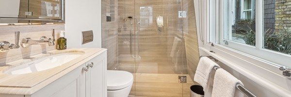 Compact bathroom with light wood finishes, glass shower, toilet, vanity, and large window with view of exterior planting.