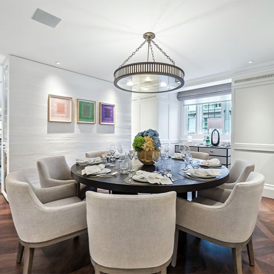 Elegant dining room with a round dark wood table set for six, surrounded by beige upholstered armchairs, a central floral arrangement, and contemporary art on the wall, under a modern chandelier.