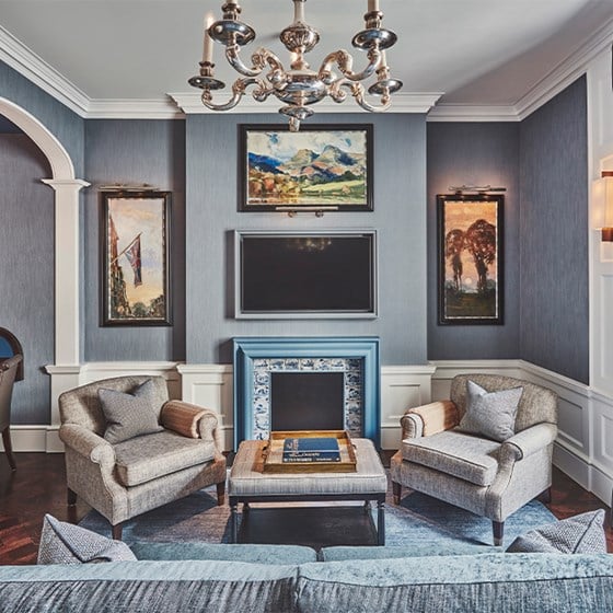 A refined, contemporary sitting room featuring plush armchairs, elegant artwork, and a stately fireplace beneath a grand chandelier, exuding timeless luxury.
