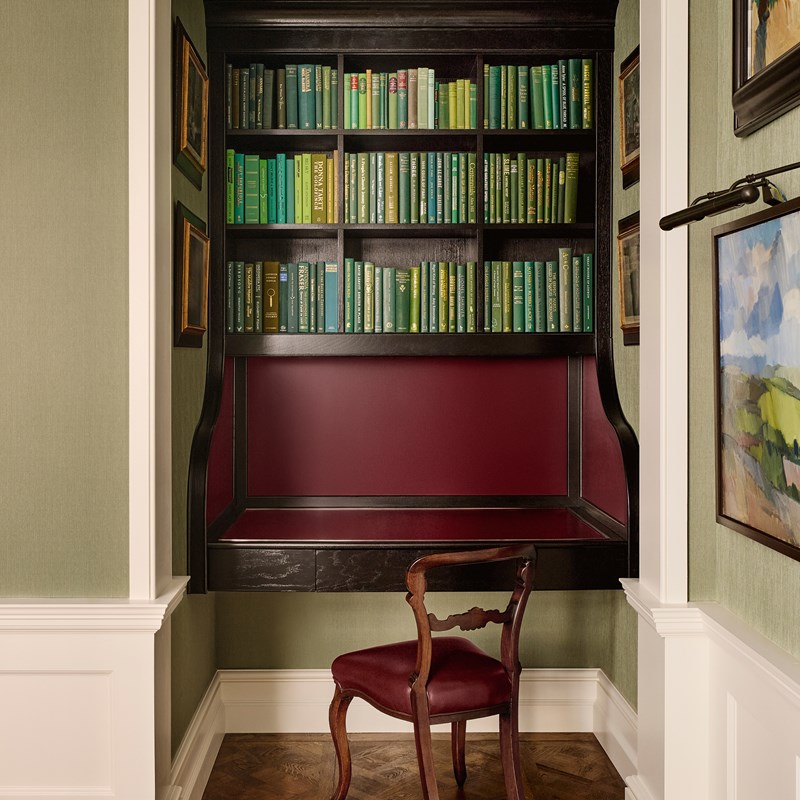 An elegant writing nook with deep burgundy panelling, a dark wood desk, and a curated bookshelf of green-bound books, framed by sage green walls and classical artwork.