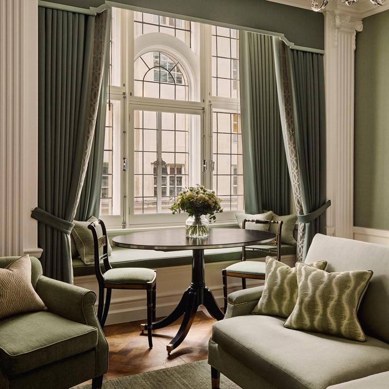 A refined sitting and dining area featuring a round black table, green upholstered seating, and grand sash windows dressed with heavy sage drapery, allowing natural light to flood the space.