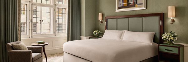 A tranquil bedroom showcasing a plush white bed with a green leather headboard, flanked by soft lighting and floral arrangements, set against sage green walls and tall period windows.