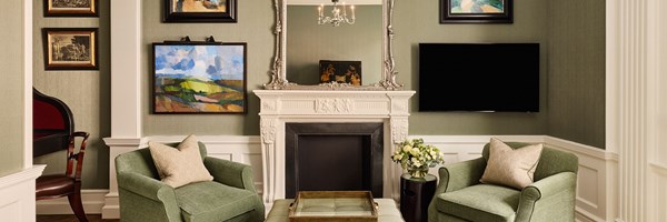 A tastefully appointed sitting room with green armchairs, a white decorative fireplace, and curated modern and traditional art pieces, blending heritage charm with contemporary elegance.