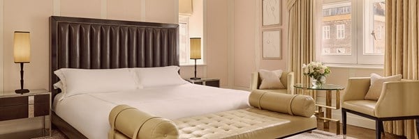 Serene bedroom with a king-sized bed, tufted leather bench, two armchairs around a glass table, and tall windows dressed with draped curtains.