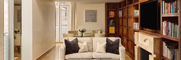 Spacious open-plan living and dining area with cream furnishings, built-in wooden bookcases, a stone fireplace, a flat-screen TV, and a glass dining table set beside French doors leading to a balcony.