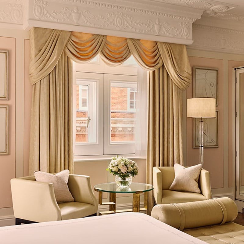 Cosy bedroom seating area with two cream armchairs, a round glass table, fresh flowers, and tall drapes framing a double window.