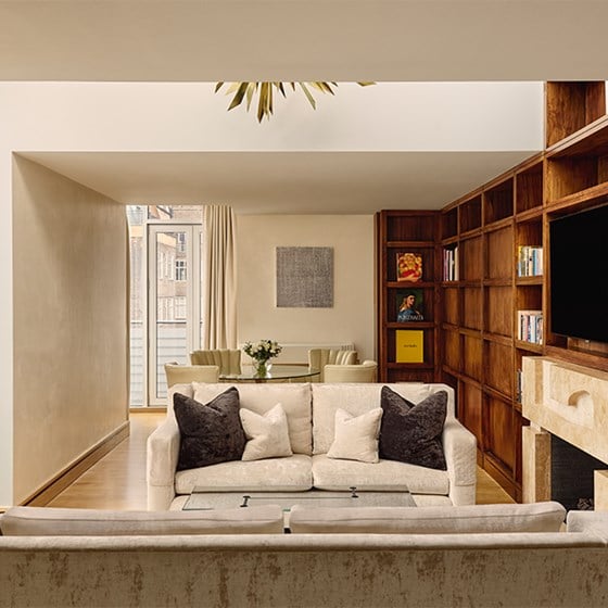 A warm-toned sitting room featuring cream sofas with dark accent cushions, a built-in wooden bookcase, a fireplace with a television above, and a dining area in the background with a round glass table and four chairs.